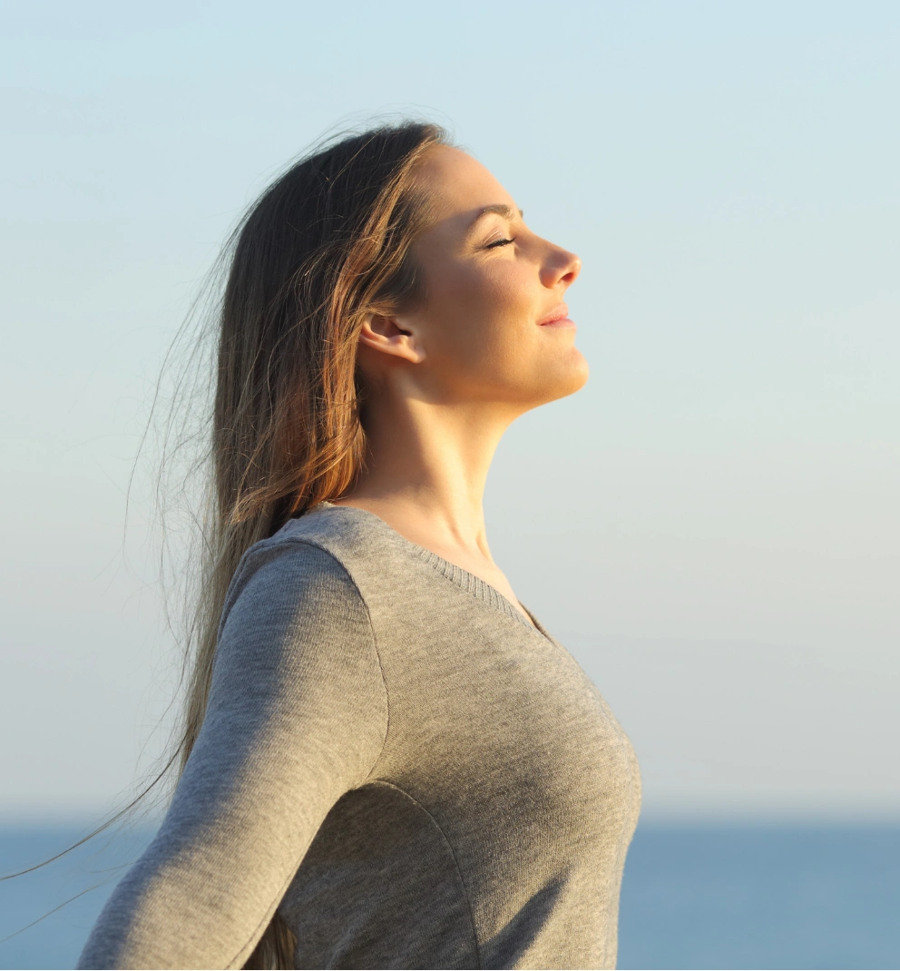 breathing Woman in a gray sweater with closed eyes, enjoying the breeze by the seaside under a clear sky.