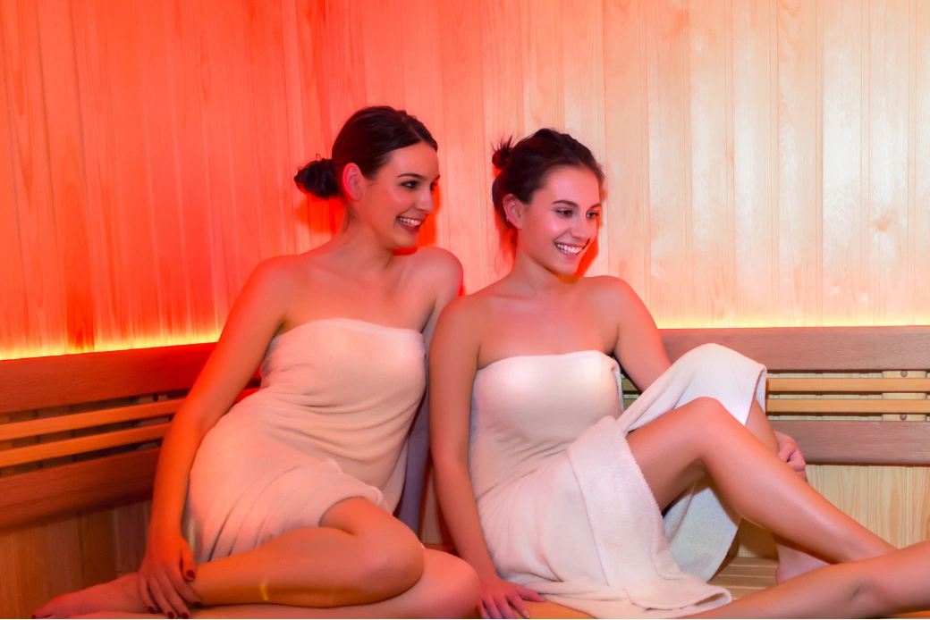 Two women in towels smiling and sitting in a warmly lit wooden sauna.