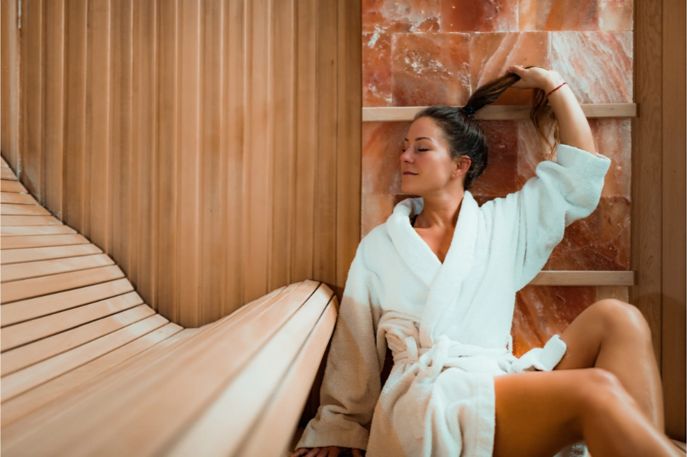 Person relaxing in a sauna, wearing a white robe, eyes closed, with hand gently holding their hair.