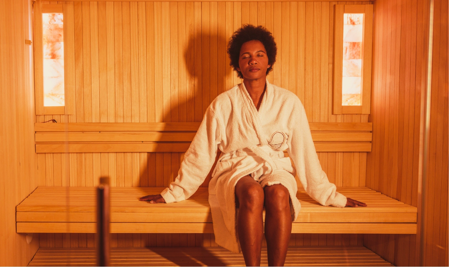 Person relaxing in a sauna, sitting on a wooden bench, wearing a white robe with eyes closed and a peaceful expression.
