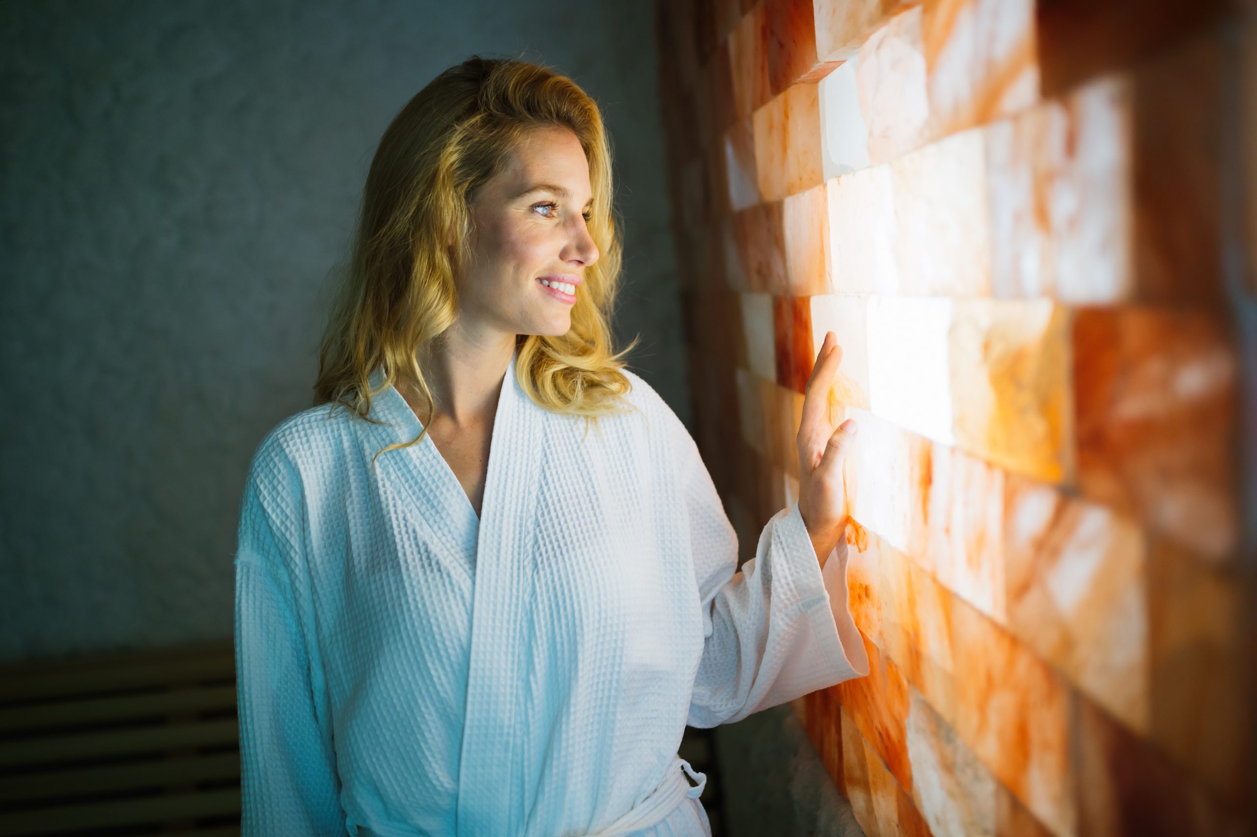 Infrared Sauna: Woman in a white robe smiling while enjoying the benefits of salt therapy, gently touching an illuminated salt wall.